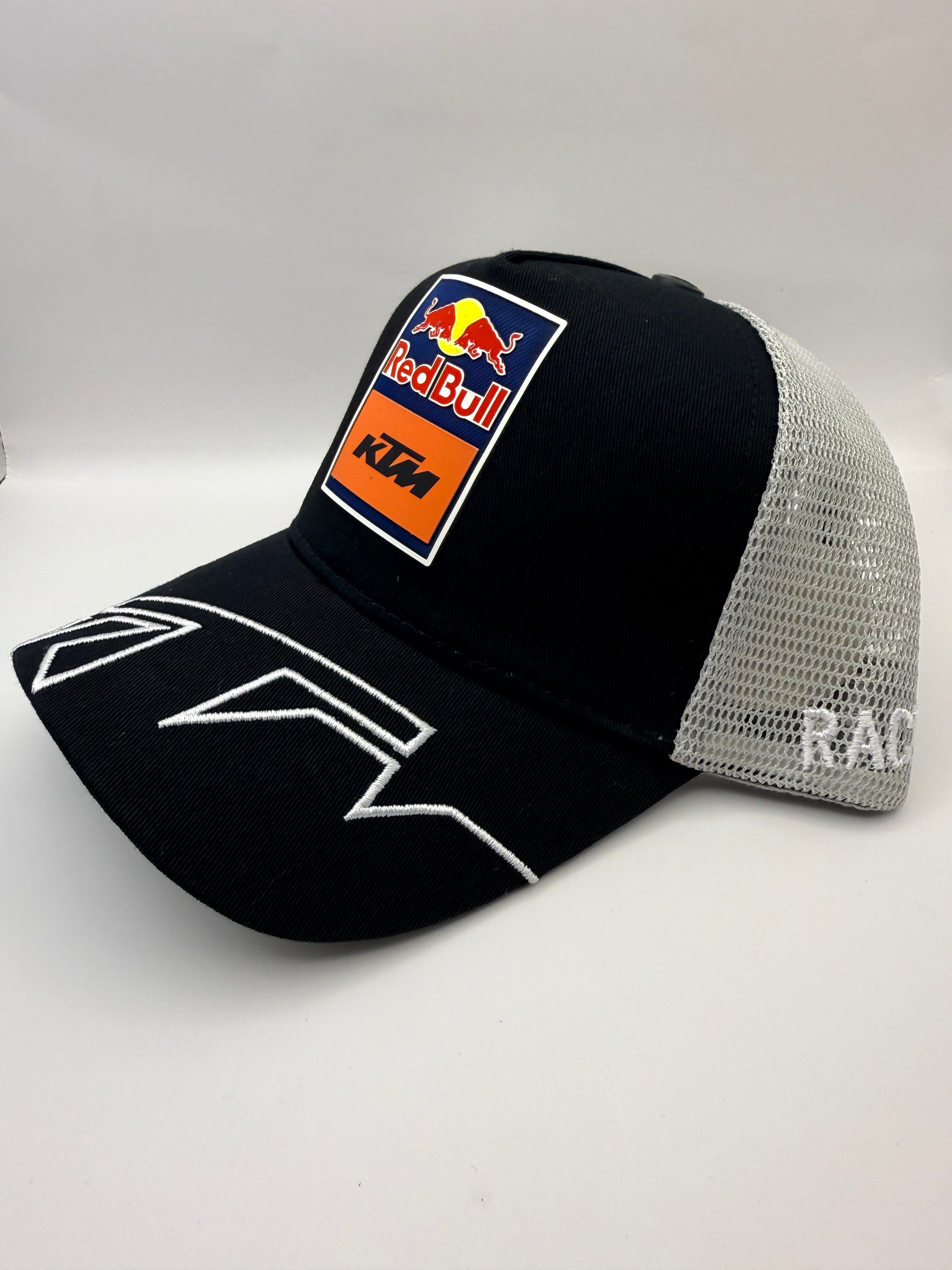 A black and silver mesh trucker hat featuring a Red Bull KTM logo patch on the front and white graphic embroidery on the brim, set against a plain light gray background.